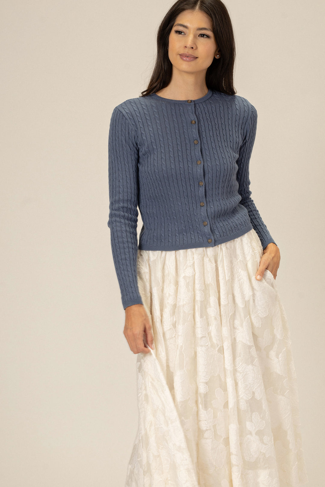 A woman wears a grey cable knit cardigan with wooden buttons, paired with a long white patterned skirt.