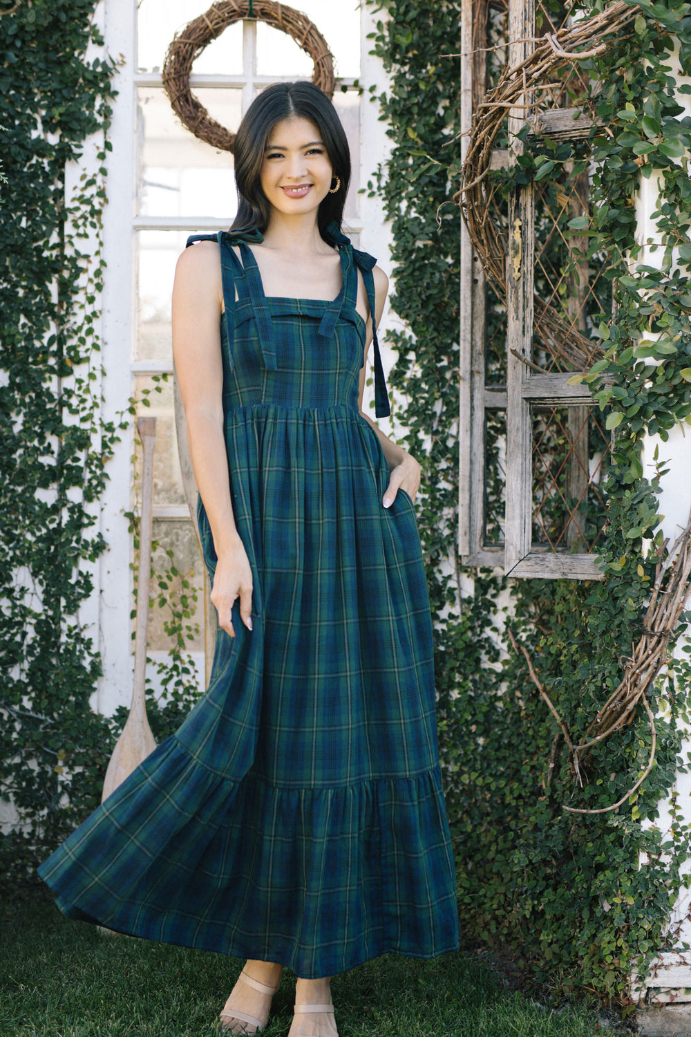 A woman in a long, flowing green and blue plaid dress stands in a garden, with a wreath-adorned window frame behind her.