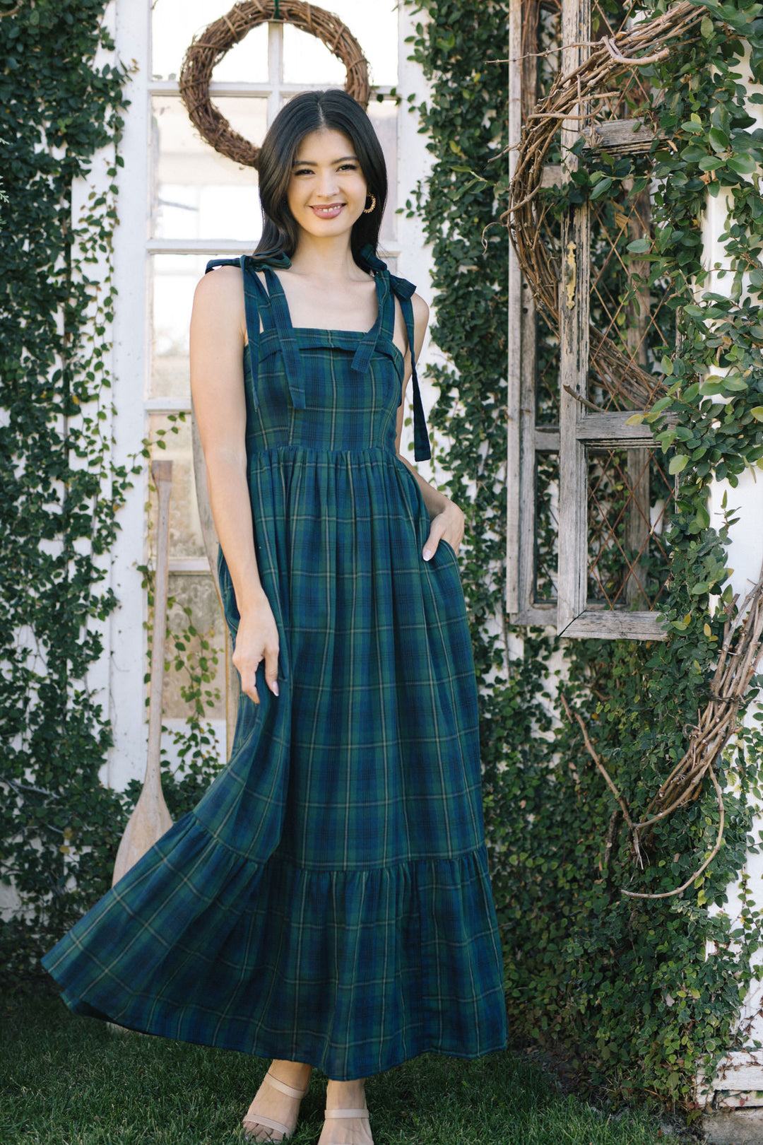A woman in a long, flowing green and blue plaid dress stands in a garden, with a wreath-adorned window frame behind her.