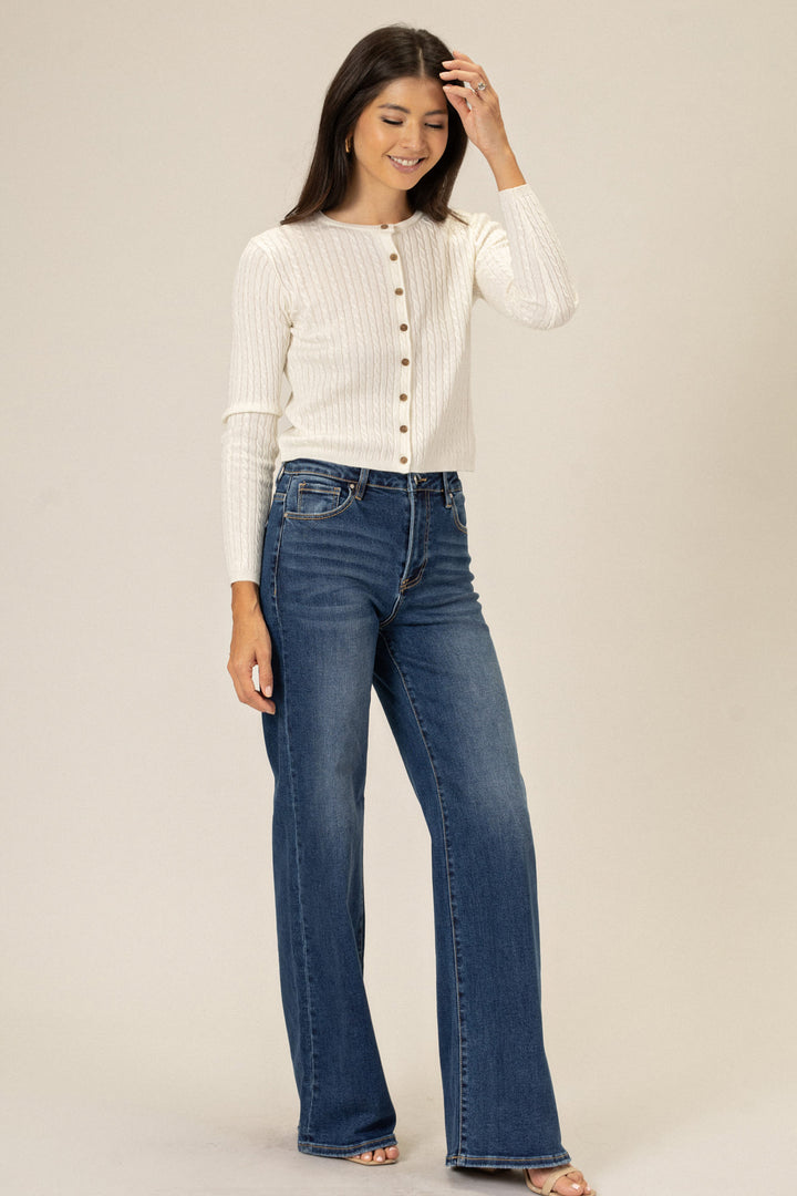 A young woman wearing a white ribbed cardigan with buttons and high-rise blue jeans, smiling and touching her hair.