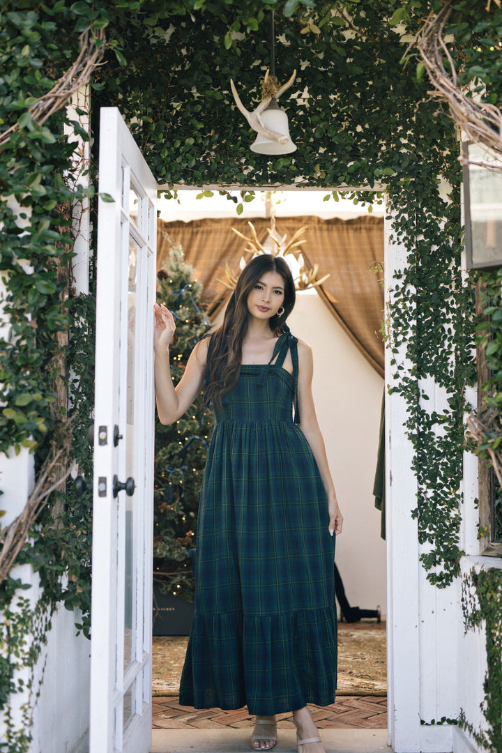 A young woman in a long, flowing green plaid dress with straps stands in a lush, verdant archway decorated with greenery.