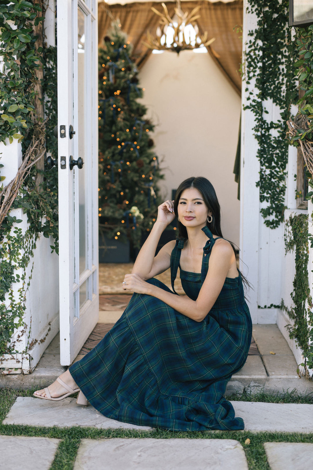 A woman stands outdoors wearing a long, plaid maxi dress in a deep green and blue hue, with a ruffled neckline and a flared, flowing skirt.