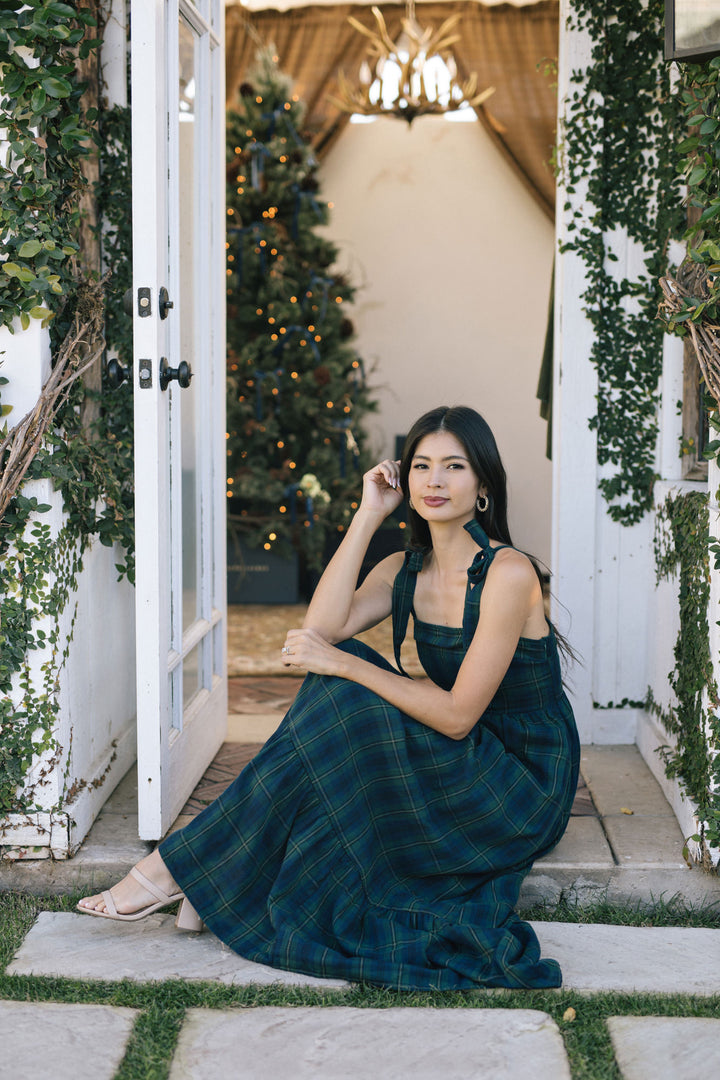 A woman stands outdoors wearing a long, plaid maxi dress in a deep green and blue hue, with a ruffled neckline and a flared, flowing skirt.