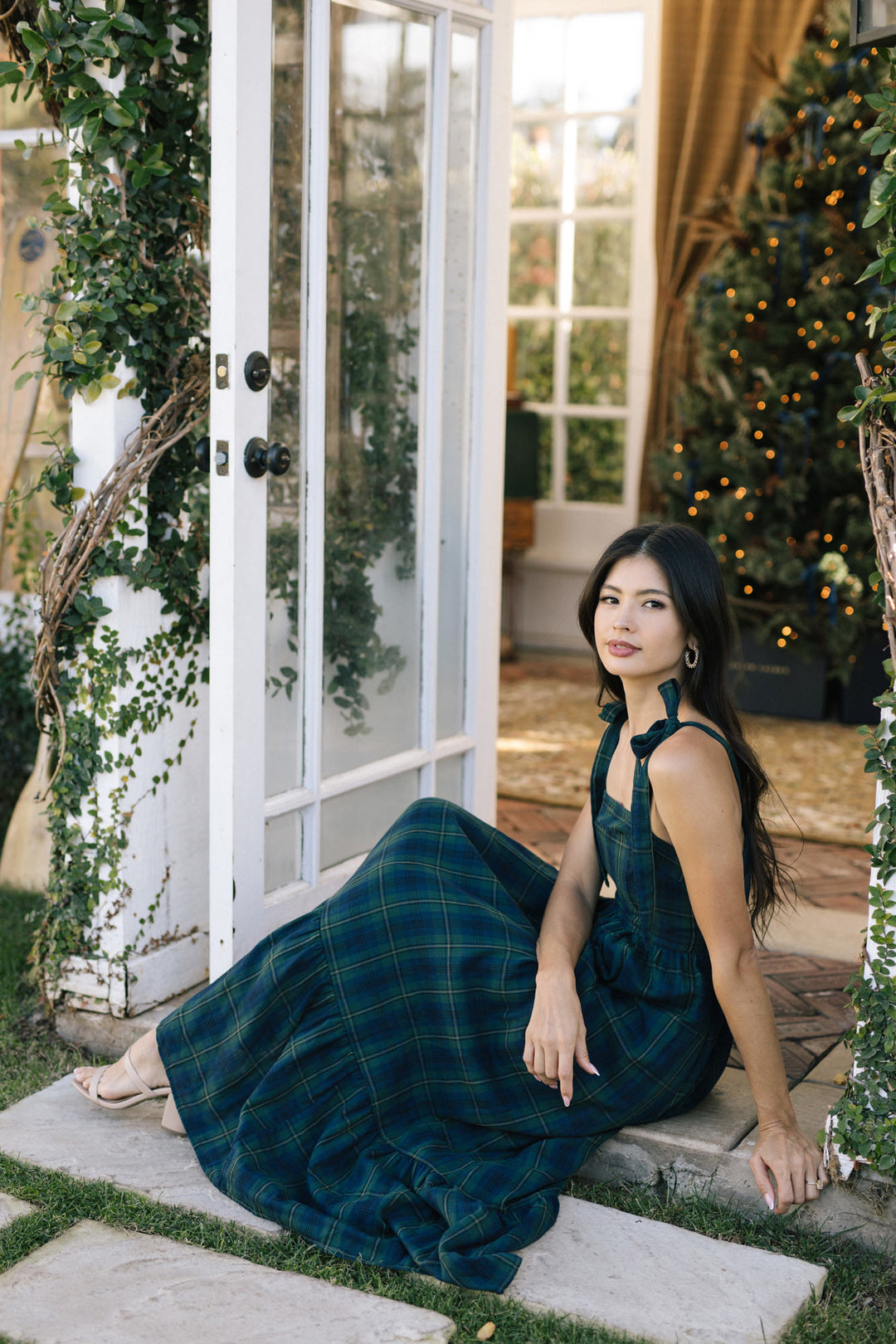 A woman wearing a floor-length, plaid dress in shades of green and navy, standing in a doorway surrounded by greenery and holiday lights.