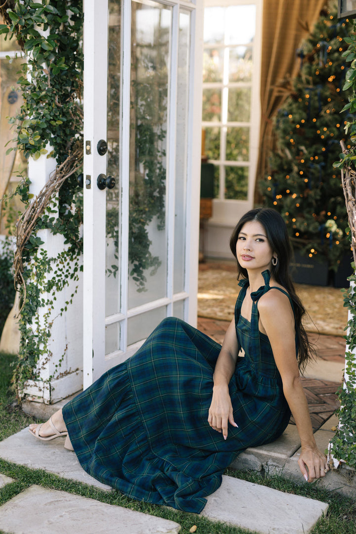 A woman wearing a floor-length, plaid dress in shades of green and navy, standing in a doorway surrounded by greenery and holiday lights.