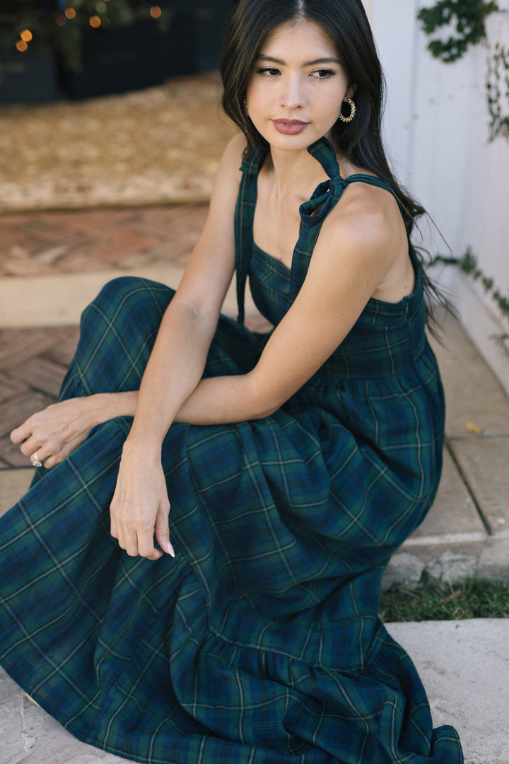 A woman wearing a long, plaid dress in shades of green and blue. The dress has a halter-style top with a bow detail and a flowy, ankle-length skirt.