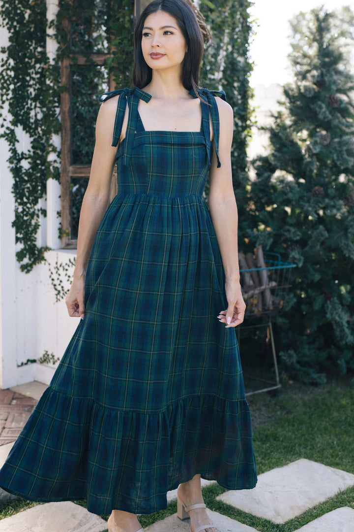 A young woman in a long, flowing green and blue plaid dress with tie shoulder straps, standing in a garden setting.