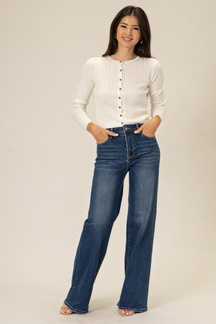 A woman wearing a white ribbed cardigan and high-rise dark wash flared jeans, standing with her hands in her pockets and smiling at the camera.