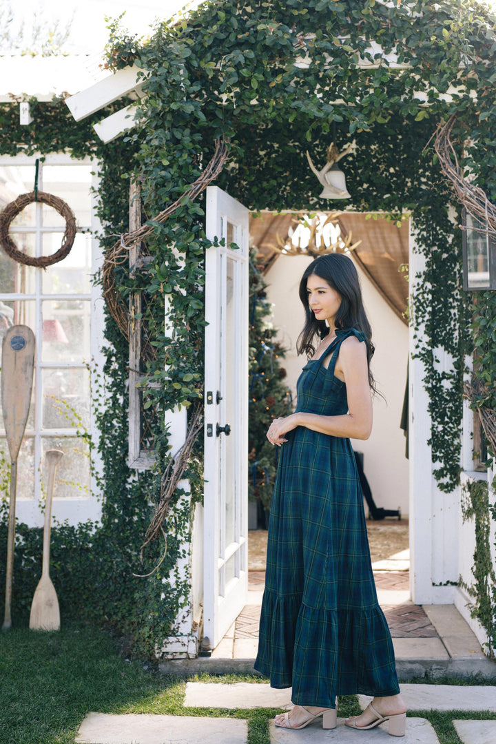 A woman standing in a floral archway wearing a long, plaid dress in a deep teal color, featuring a sleeveless halter neckline and a tiered, ruffled skirt.