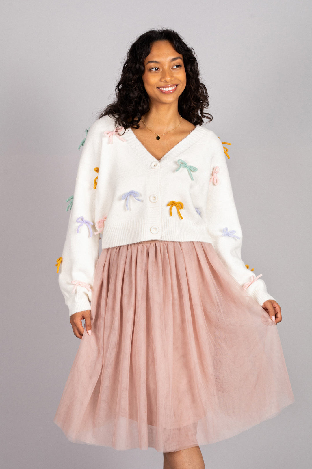 A smiling woman wearing a white cardigan with colorful bow and flower designs, paired with a pink tulle skirt.