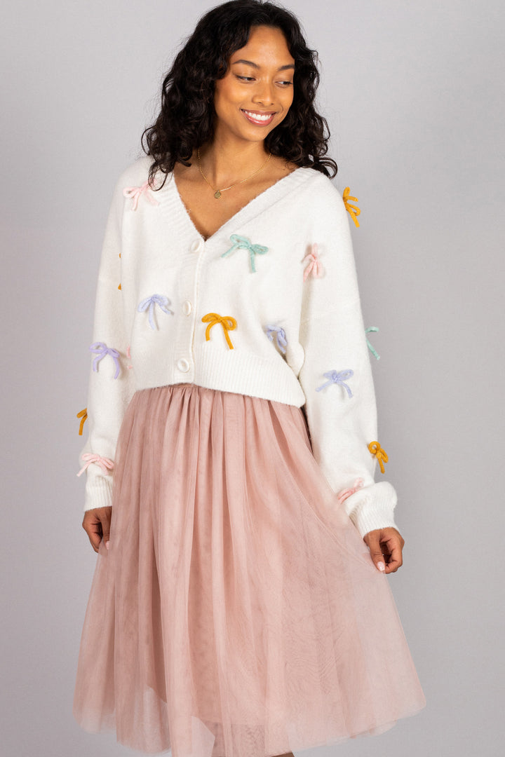 A white knit cardigan with colorful bow appliqués, paired with a soft pink tulle midi skirt, modeled by a smiling woman with curly dark hair.
