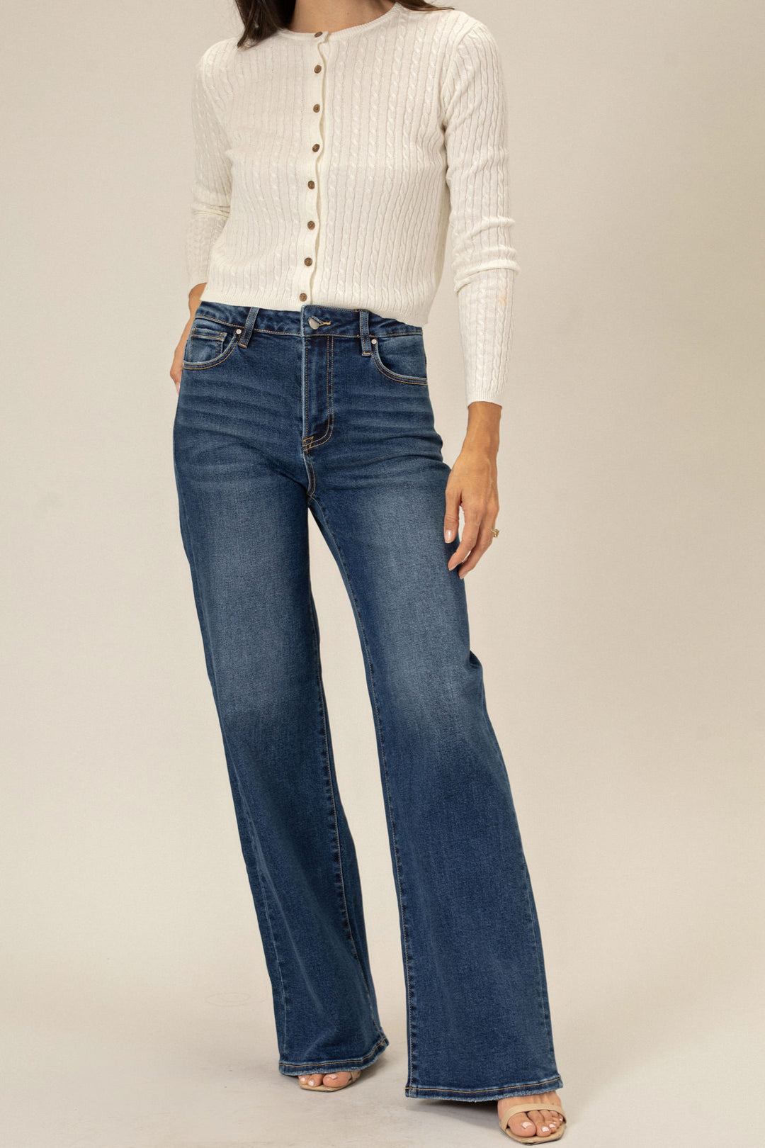 A model wearing a pair of high-waisted flared dark wash jeans and a white ribbed cardigan, standing in a neutral setting.