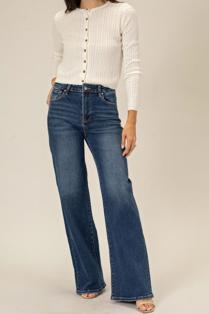A model wearing a pair of high-waisted flared dark wash jeans and a white ribbed cardigan, standing in a neutral setting.