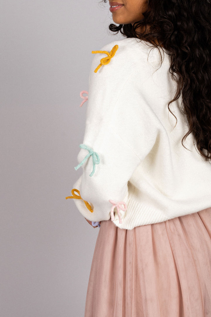 A cozy white knit cardigan with colorful bows and ties, perfect for layering over a soft pink tulle skirt.