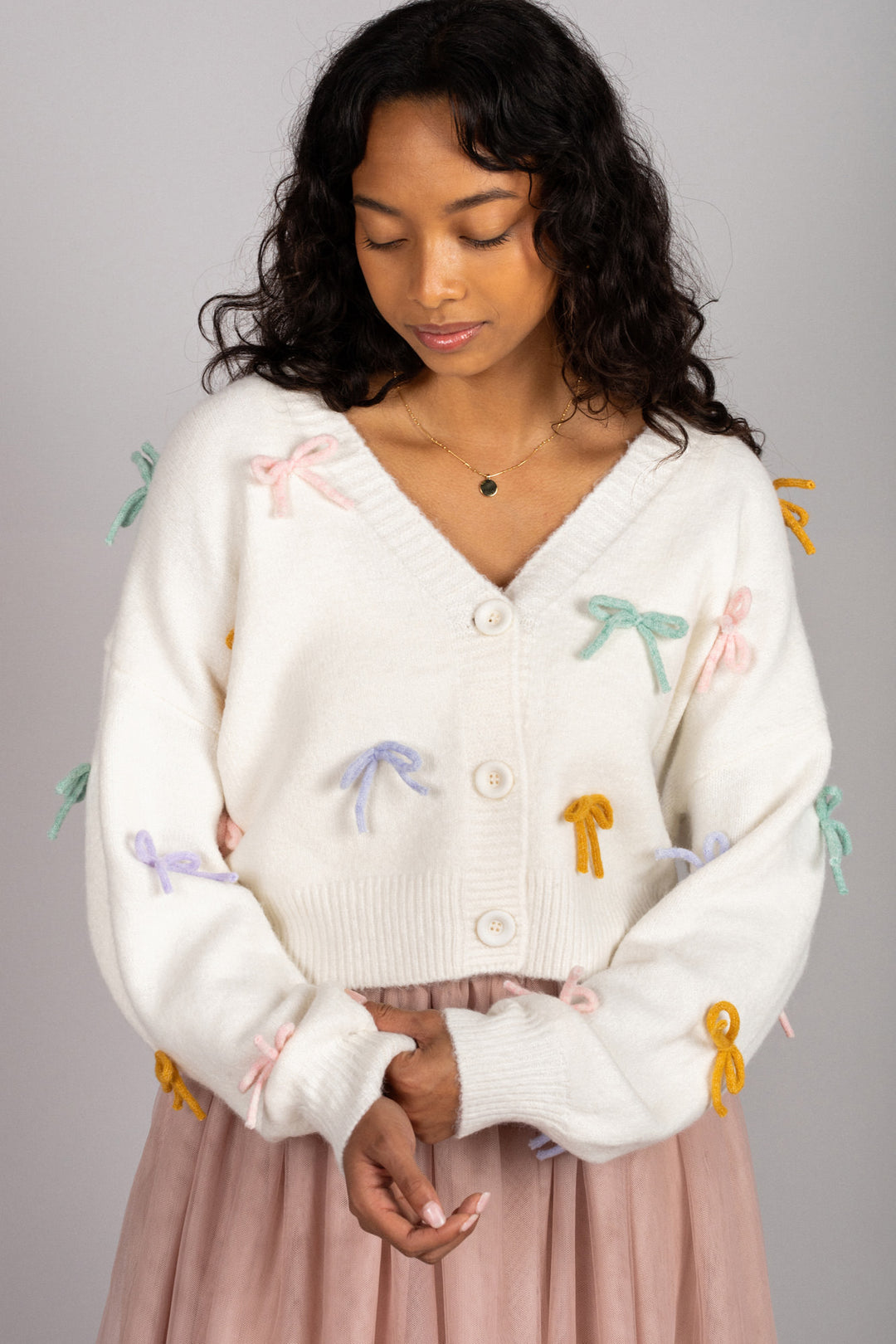 A cozy, white cardigan with colorful bow appliques, modeled by a woman with curly dark hair, wearing a blush-colored midi skirt.