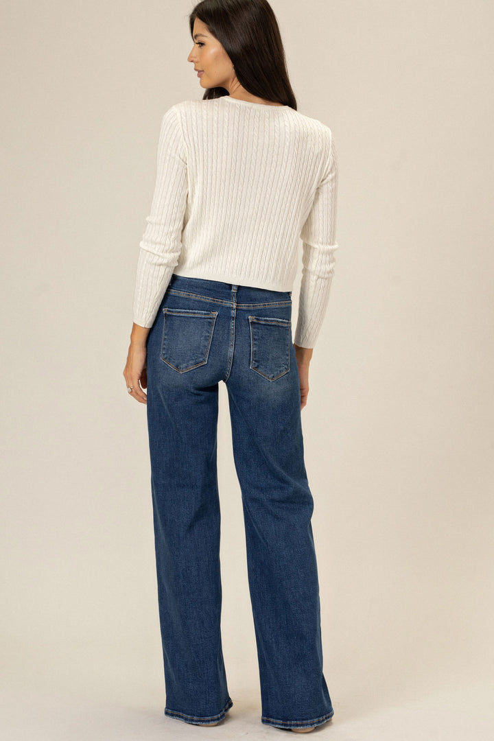 A woman wearing a white cable knit sweater and high-rise flared dark wash jeans, facing away from the camera.