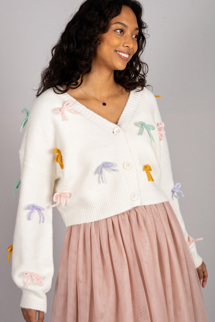 A white knitted cardigan with pastel-colored bows embellished throughout, worn by a smiling model with dark curly hair.