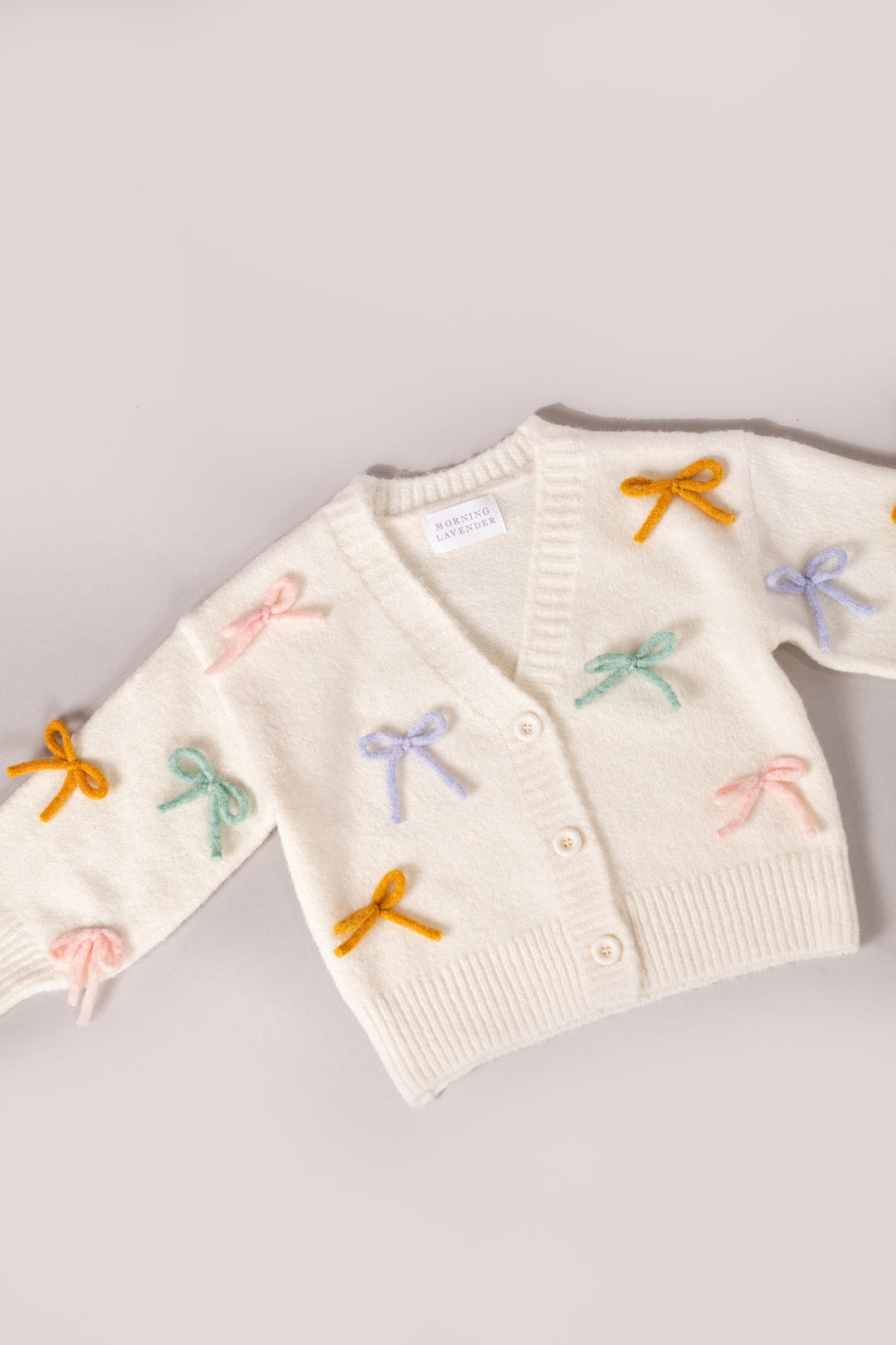 A cozy white knit cardigan with colorful bow appliques, perfect for girls' casual or dressy outfits.