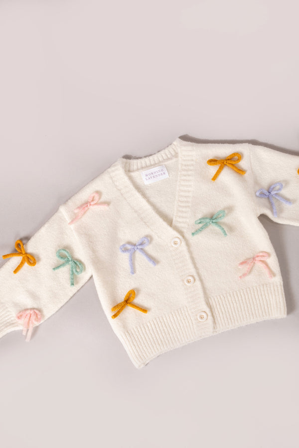 A cozy white knit cardigan with colorful bow appliques, perfect for girls' casual or dressy outfits.
