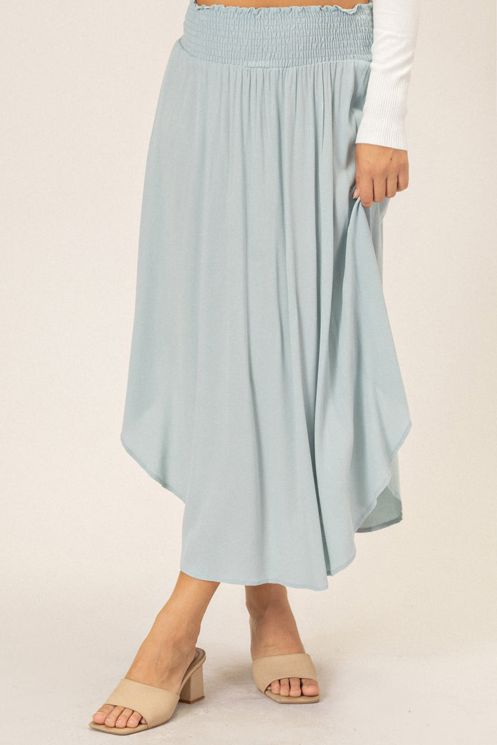 A long, light blue, flowy midi skirt with a gathered waistline and a round hem. The skirt is paired with beige slide sandals.