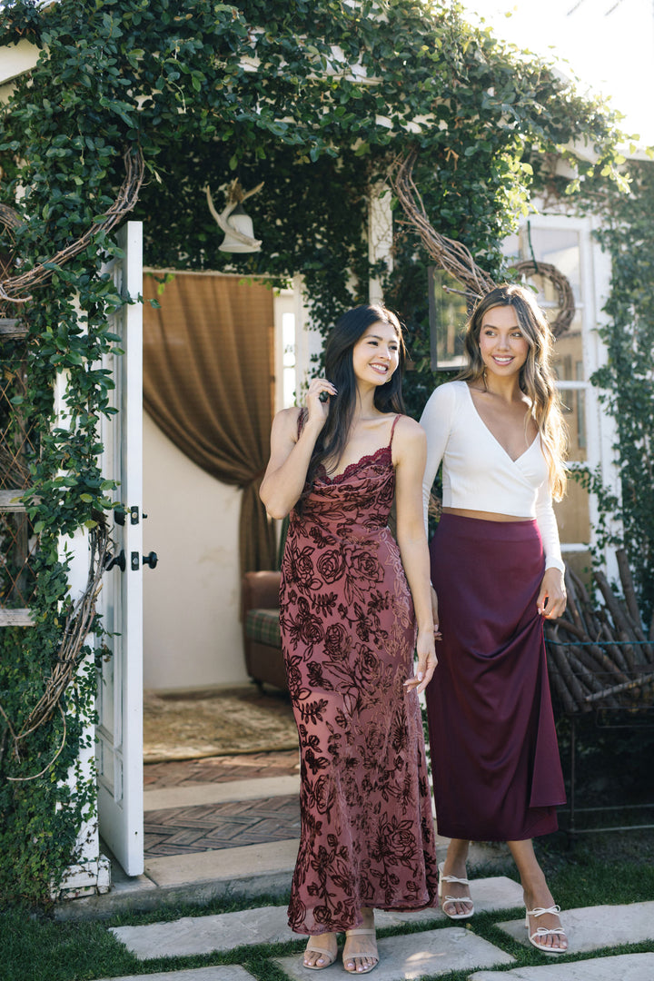 Two stylish young women posing in floral-patterned midi dress and burgundy skirt, standing in a lush, greenery-filled outdoor setting.