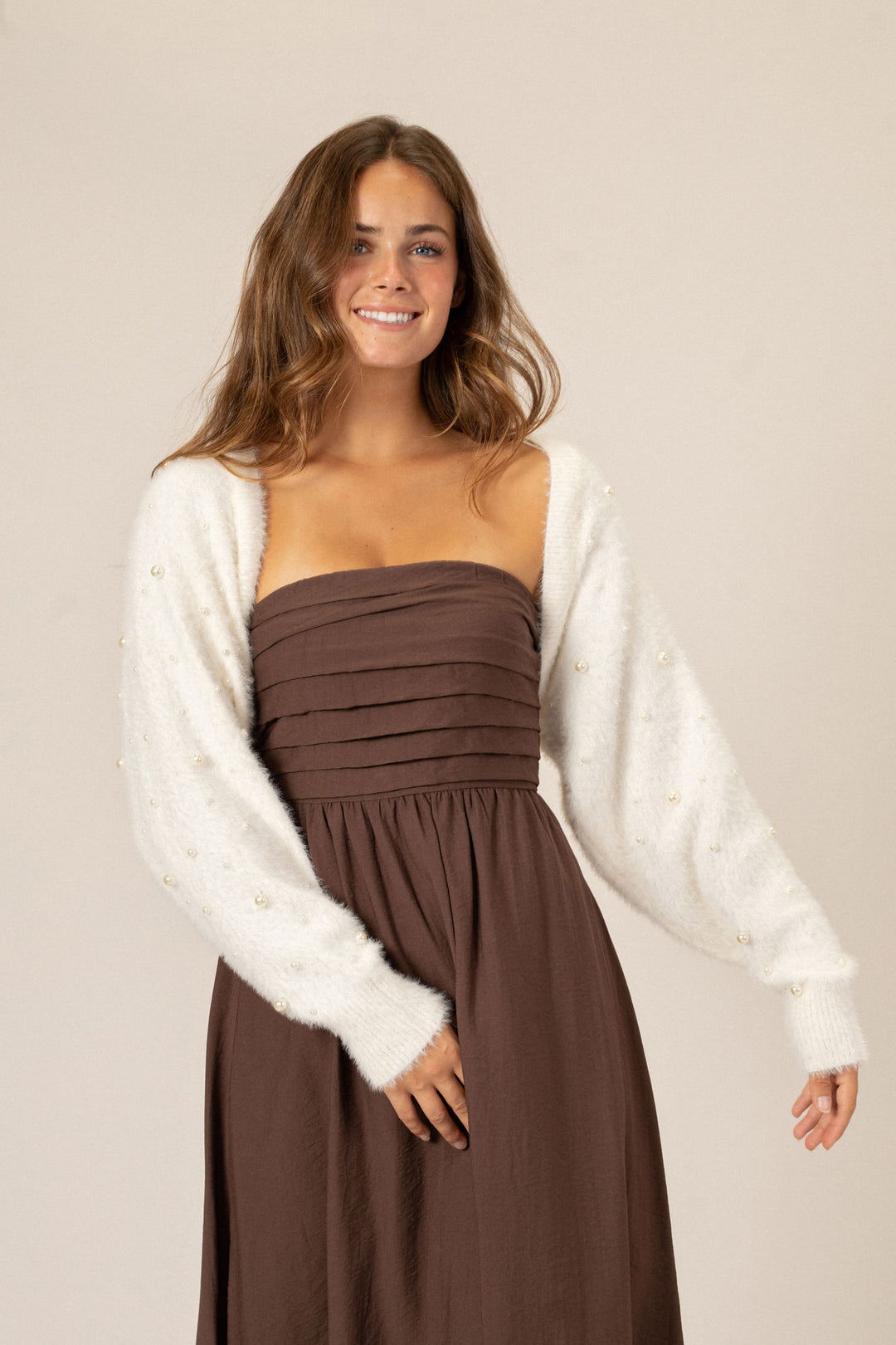 The image shows a young woman with long, wavy brown hair smiling while wearing a pleated, brown midi dress and a white fuzzy cardigan.