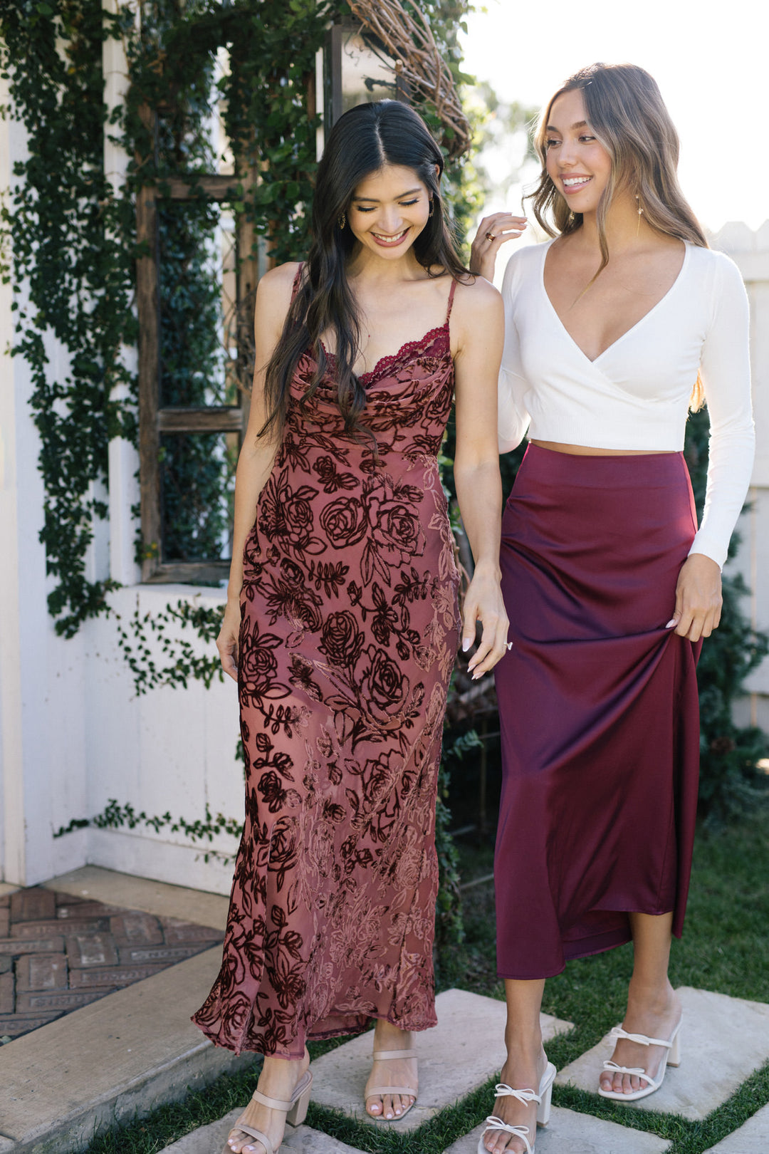 Two young women posing in colorful floral dresses and skirts, one in a burgundy midi skirt and white wrap top, the other in a burgundy floral midi dress.