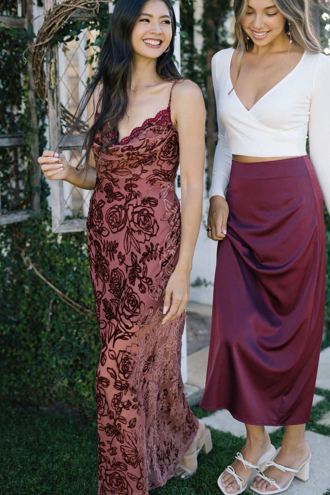 Two women stand in a garden, one wearing a burgundy floral slip dress and the other a white cropped top with a burgundy midi skirt.