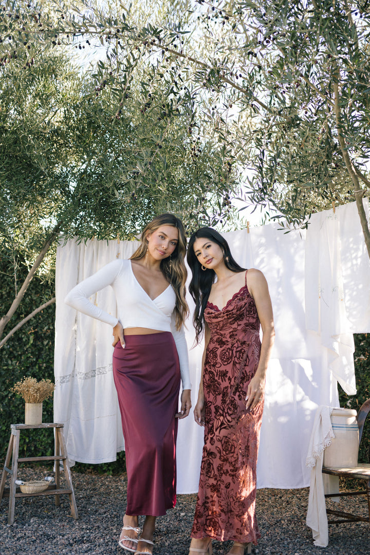 Two young women in fashionable evening attire posing against a backdrop of olive trees and white curtains.
