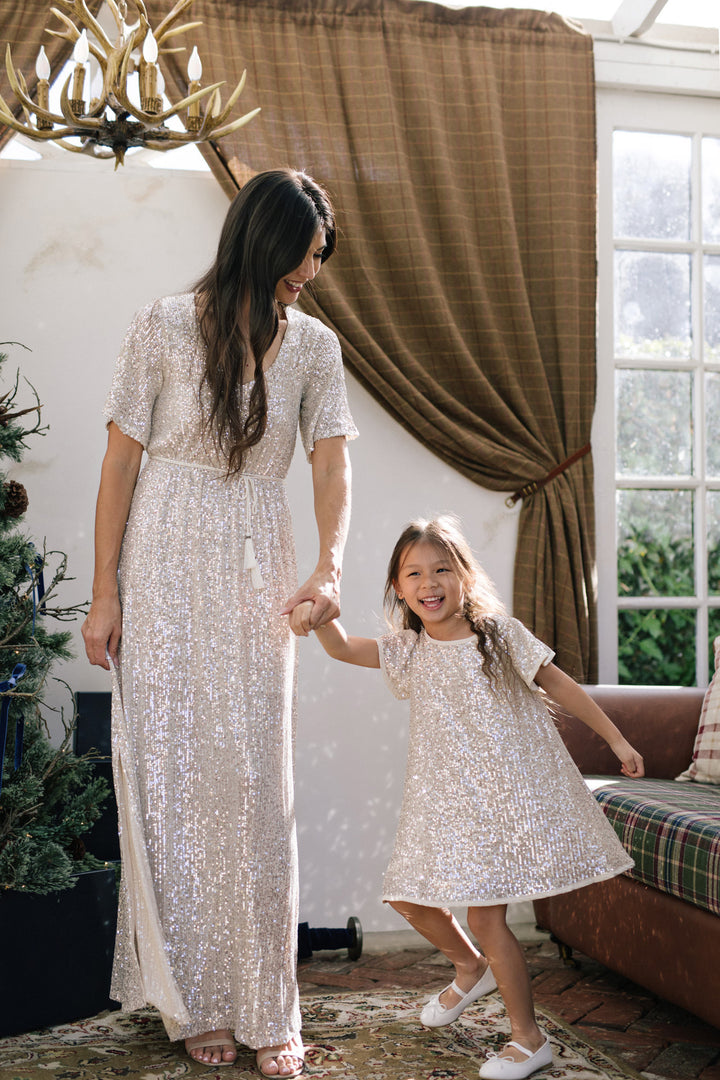 A long, shimmering silver sequin dress with a high neck, short sleeves, and a flowing maxi-length skirt, paired with a sequin-embellished child's dress in a matching style.