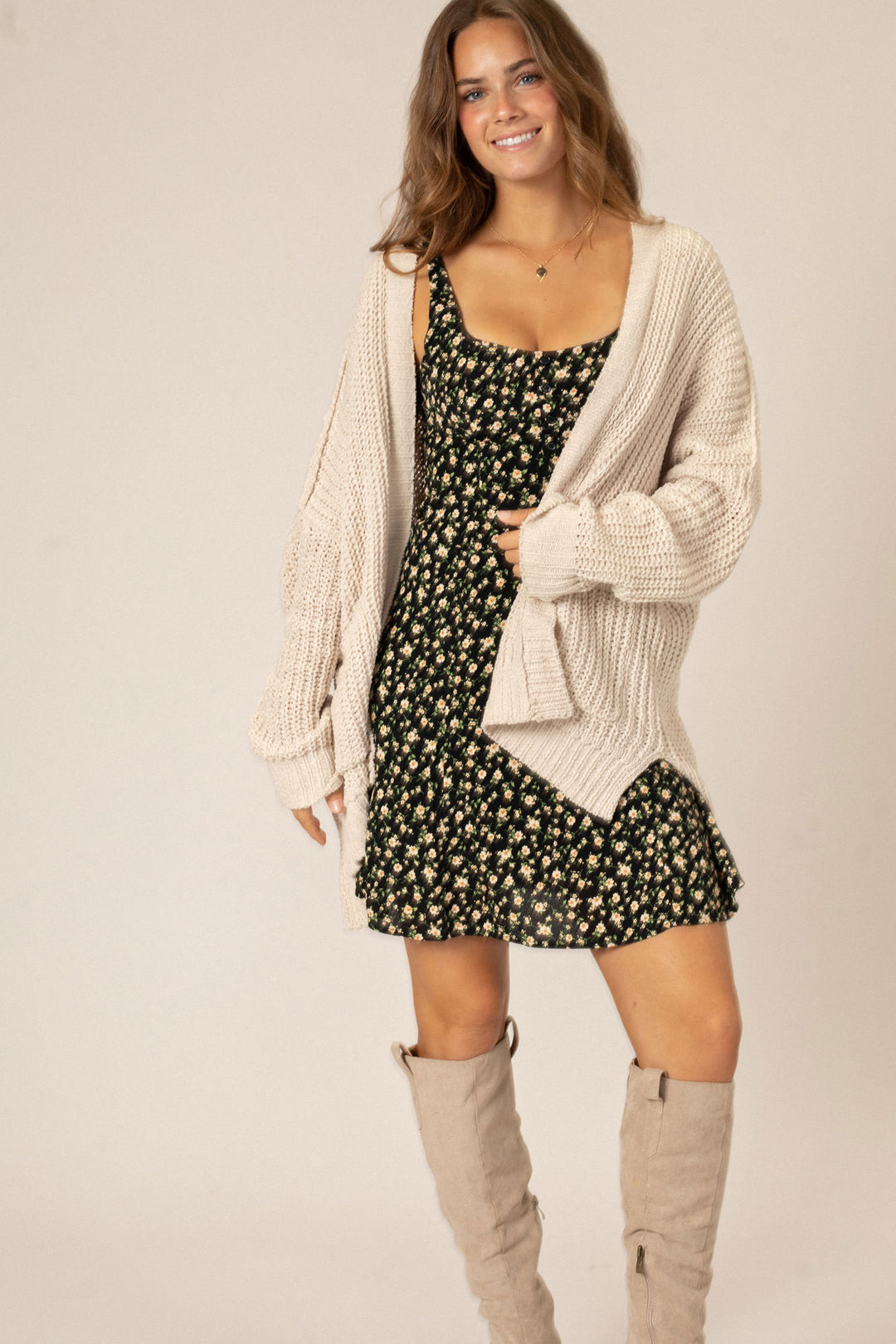 A young woman wearing a beige knit cardigan over a black floral print dress, paired with beige knee-high boots, standing and smiling against a light background.