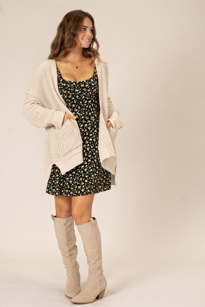 A smiling woman in a black floral dress with a long tan cardigan and beige knee-high boots, showcasing a comfy, stylish ensemble.