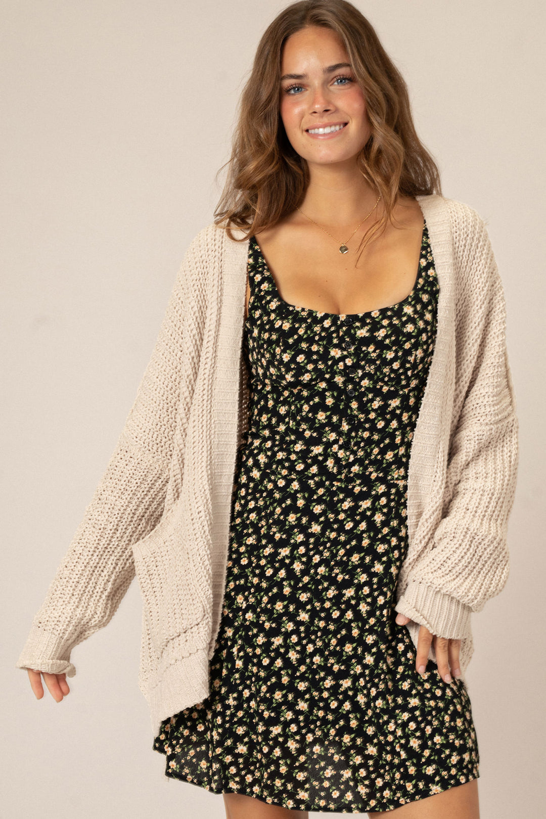 A smiling woman wearing a floral print dress and a cozy beige knit cardigan, posing against a neutral background.