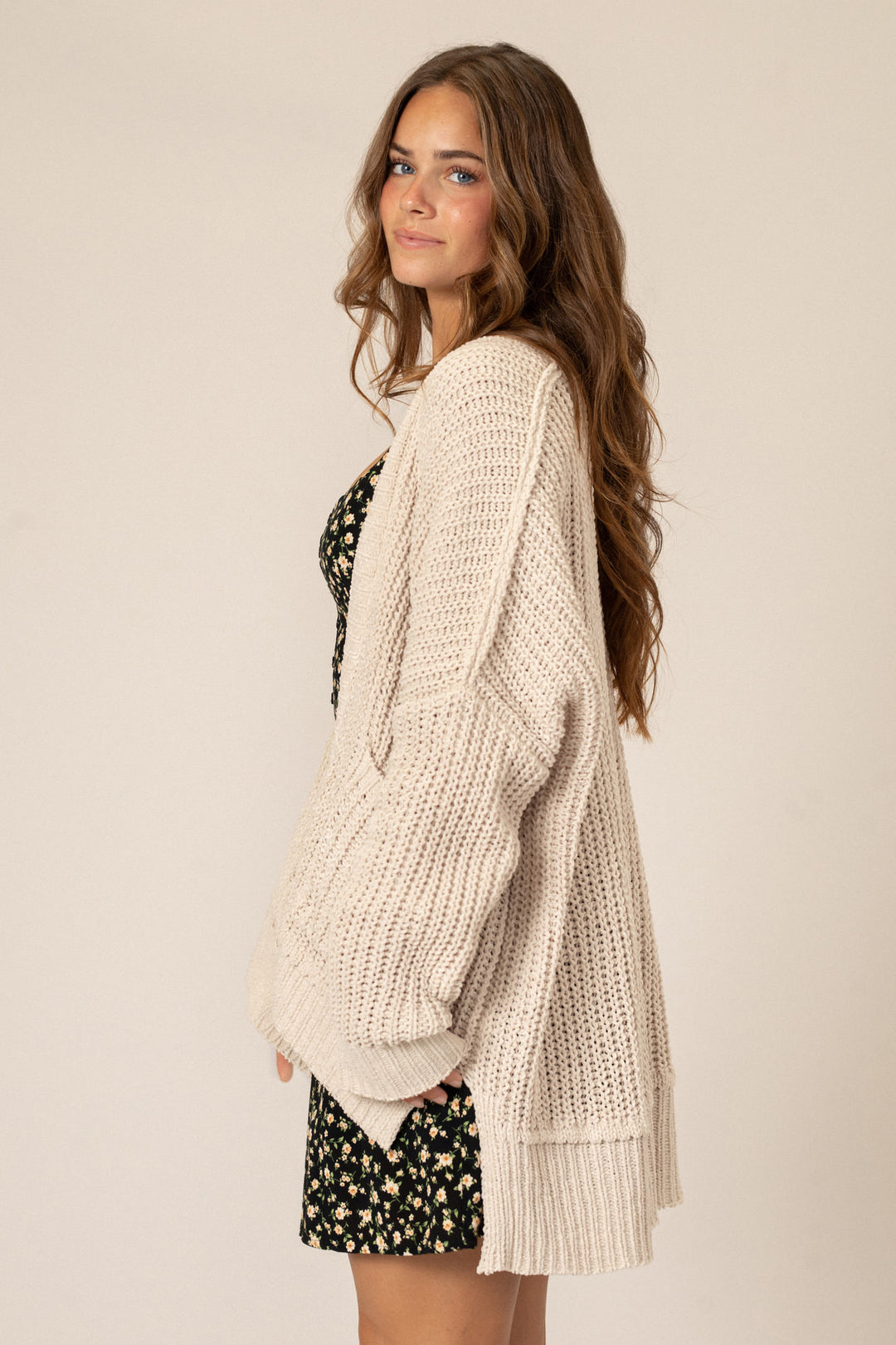 A cozy, ivory-colored knit cardigan with a relaxed, open silhouette and long sleeves, worn over a floral print dress.