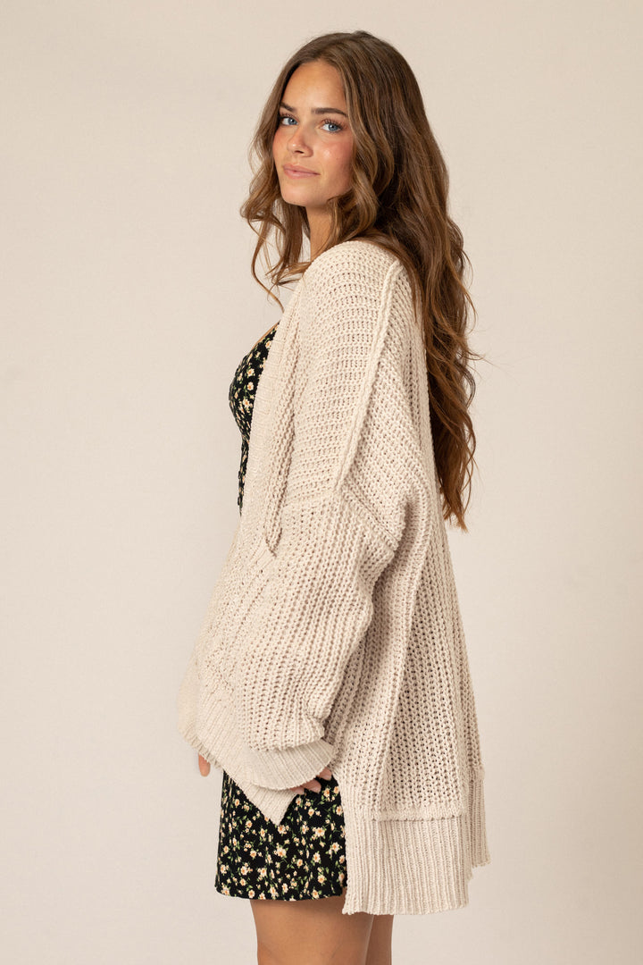 A cozy, ivory-colored knit cardigan with a relaxed, open silhouette and long sleeves, worn over a floral print dress.
