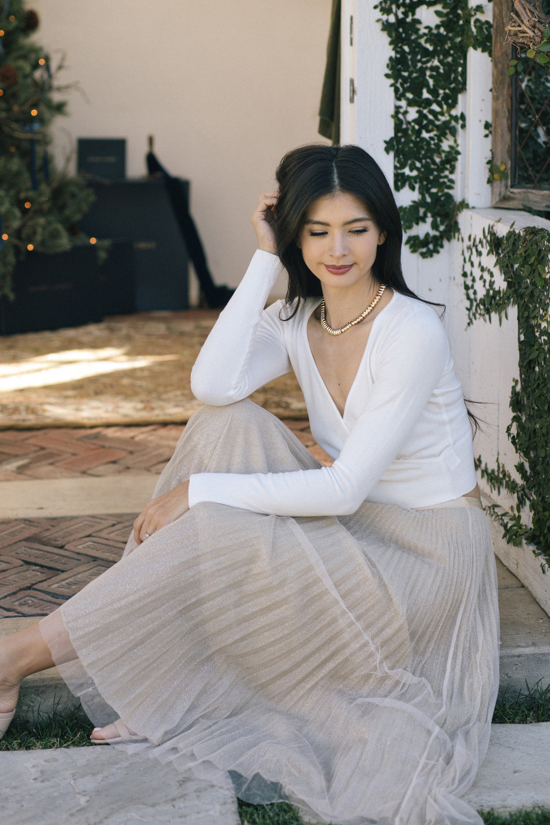 A young woman in a white blouse and long pleated skirt sits on a paved surface surrounded by greenery, with a necklace of round beads.