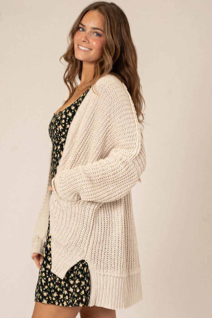 A smiling woman wearing a cozy cream-colored knit cardigan with a relaxed, draped silhouette over a black and white floral mini dress.