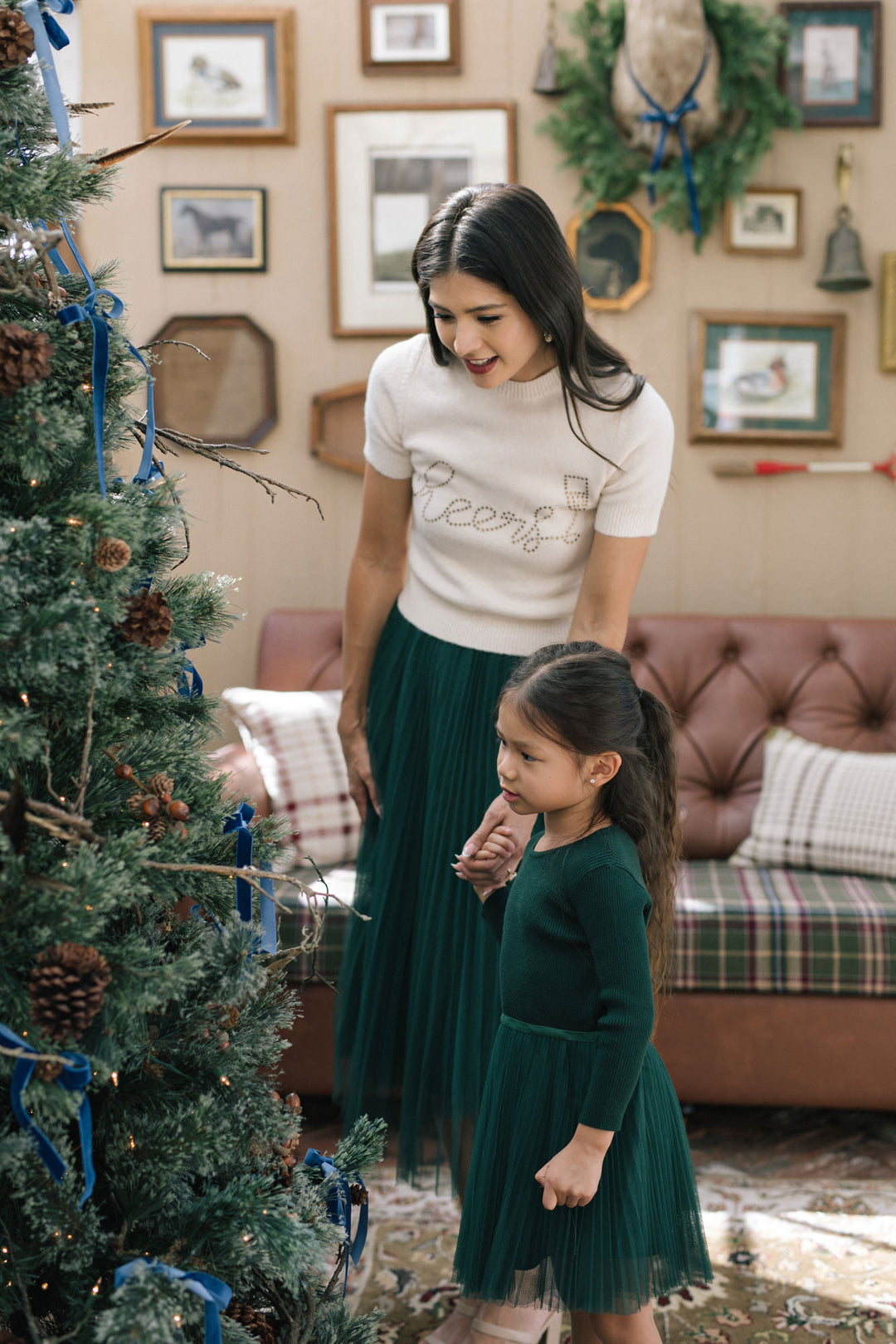 A young girl in a green pleated tulle dress stands beside a decorated Christmas tree, smiling at the camera.