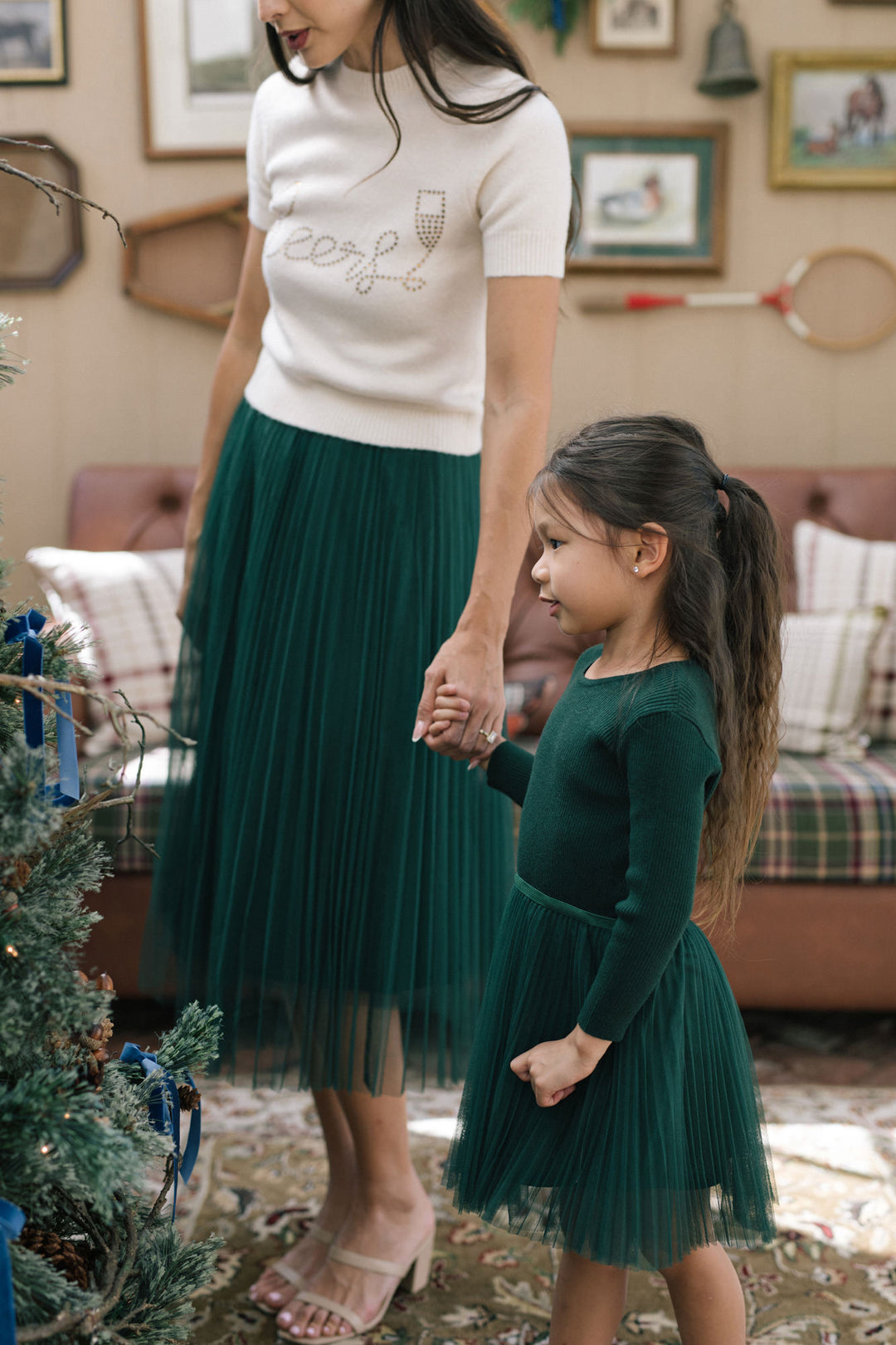 A young girl wearing a green, pleated tulle dress stands next to her mother in a cozy, holiday-decorated room.