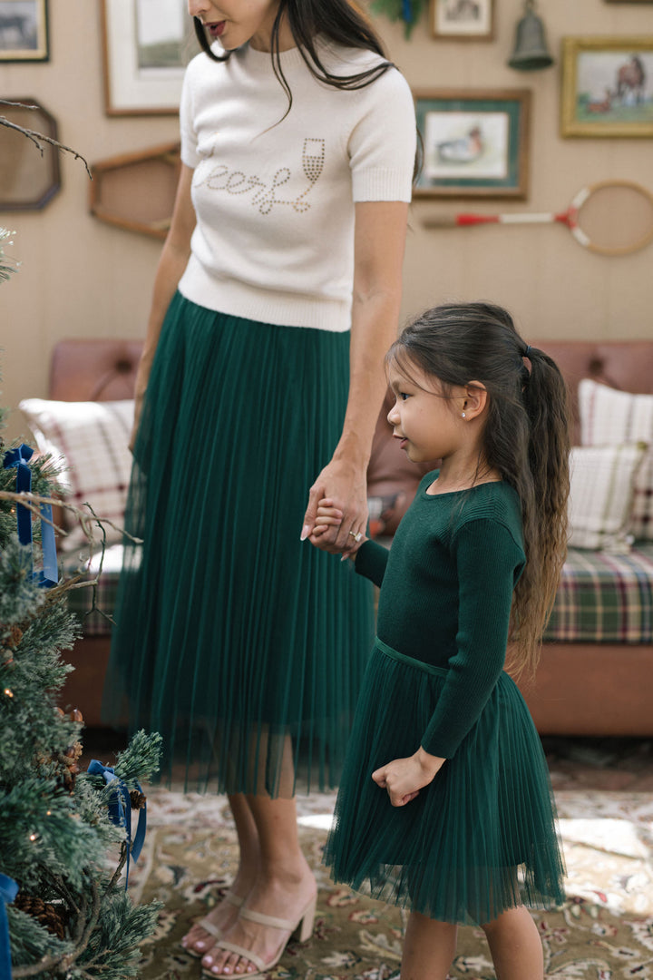 A young girl wearing a green, pleated tulle dress stands next to her mother in a cozy, holiday-decorated room.