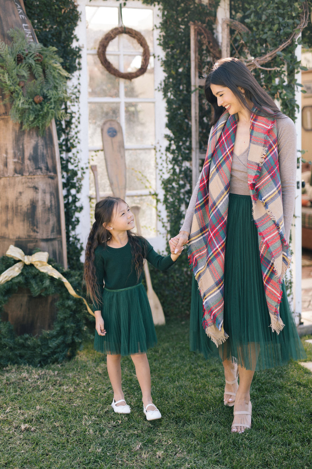 A young girl wearing a green tulle dress stands hand-in-hand with a woman wearing a colorful plaid scarf, both smiling in a lush outdoor setting.