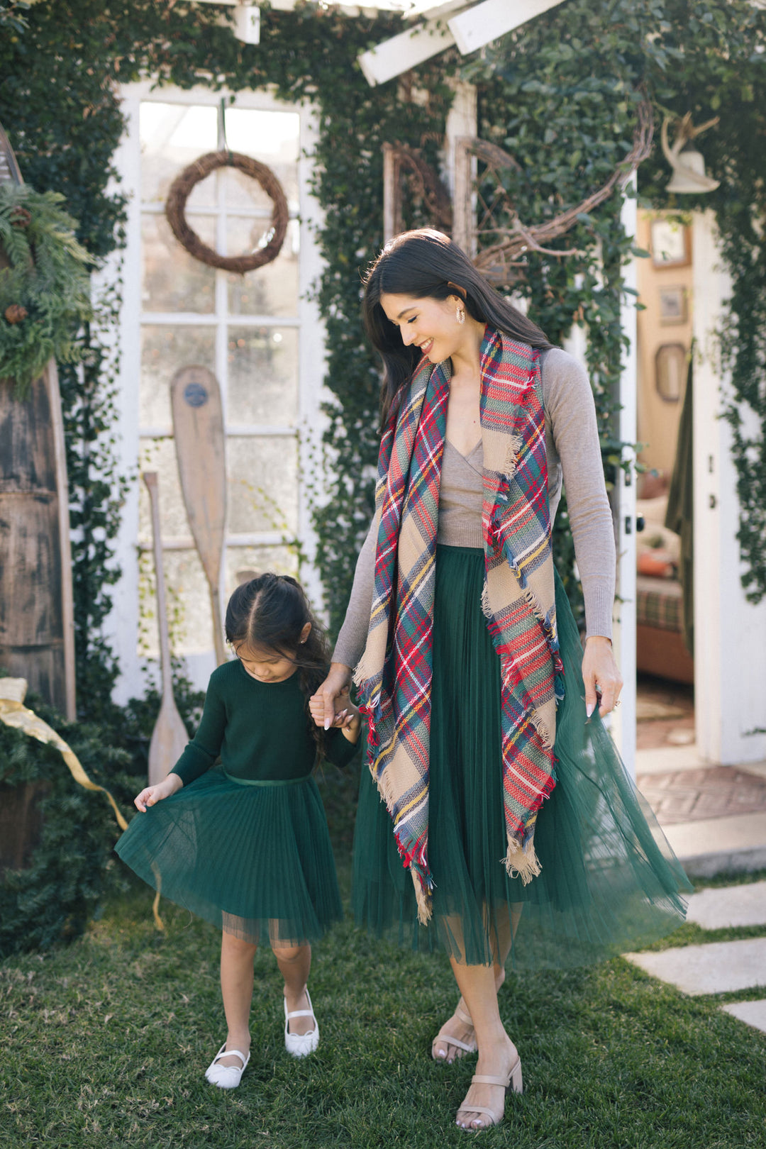 A woman in a colorful plaid scarf and long green tulle skirt stands with a young girl in a matching green dress, both smiling in a garden setting with a vintage-style window frame.