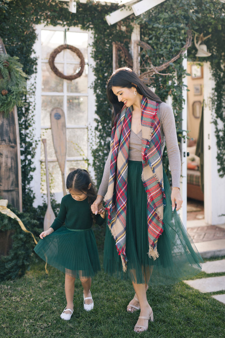 A woman in a colorful plaid scarf and long green tulle skirt stands with a young girl in a matching green dress, both smiling in a garden setting with a vintage-style window frame.