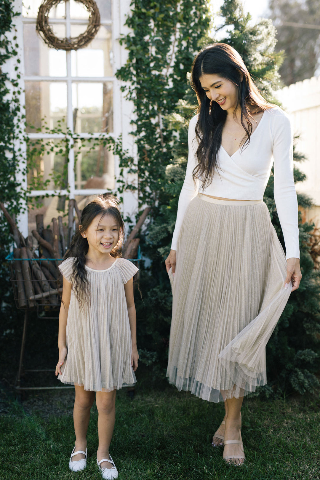 Two smiling young girls, one wearing a long pleated cream-colored dress and the other wearing a pleated cream-colored knee-length dress, standing together in a lush outdoor setting with a window frame and wreath visible.
