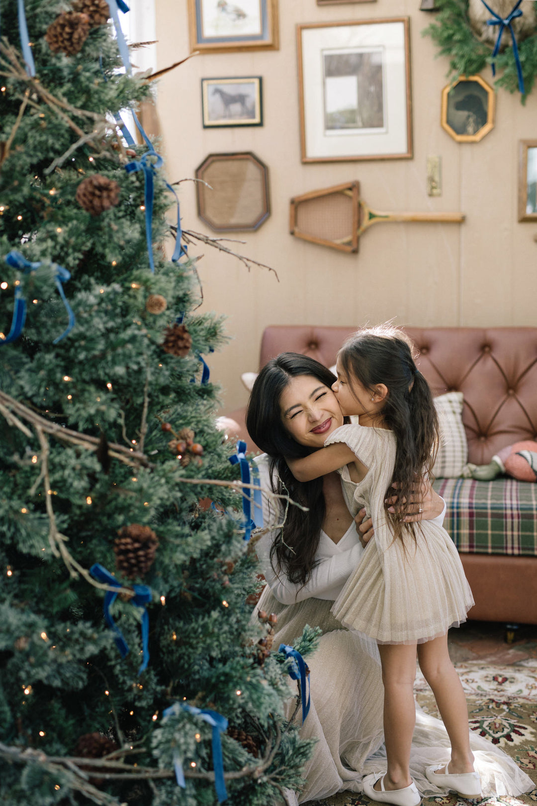 Two young girls hugging in front of a Christmas tree, with the older girl in a white pleated dress and the younger girl in a tulle skirt, surrounded by festive decor.