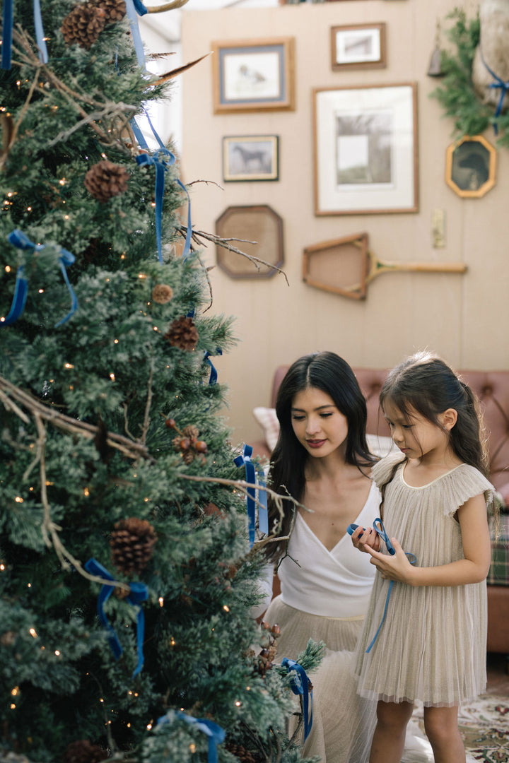 Two young girls, one wearing a white dress and the other a pleated beige dress, standing in front of a Christmas tree with gold ornaments and family photos on the wall.