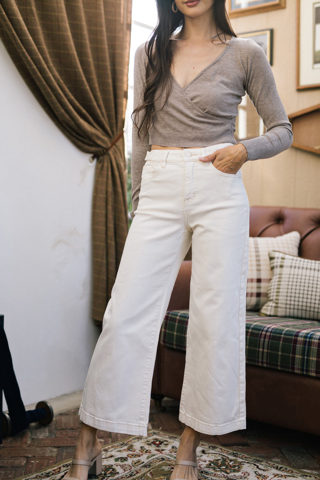 Beige cropped wide-leg jeans with a wrap-style knit top and long sleeves, set against a cozy interior backdrop.