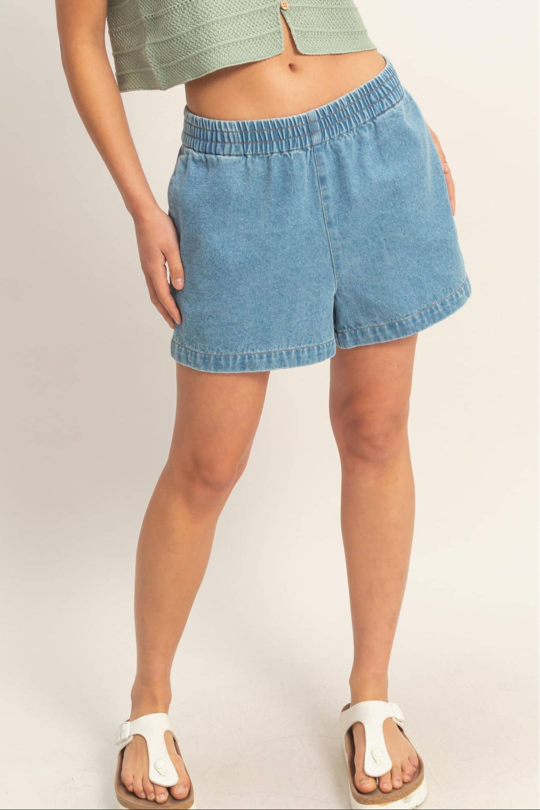 Light blue denim shorts with an elastic waistband and ruffled trim, styled with a green cropped tank top.