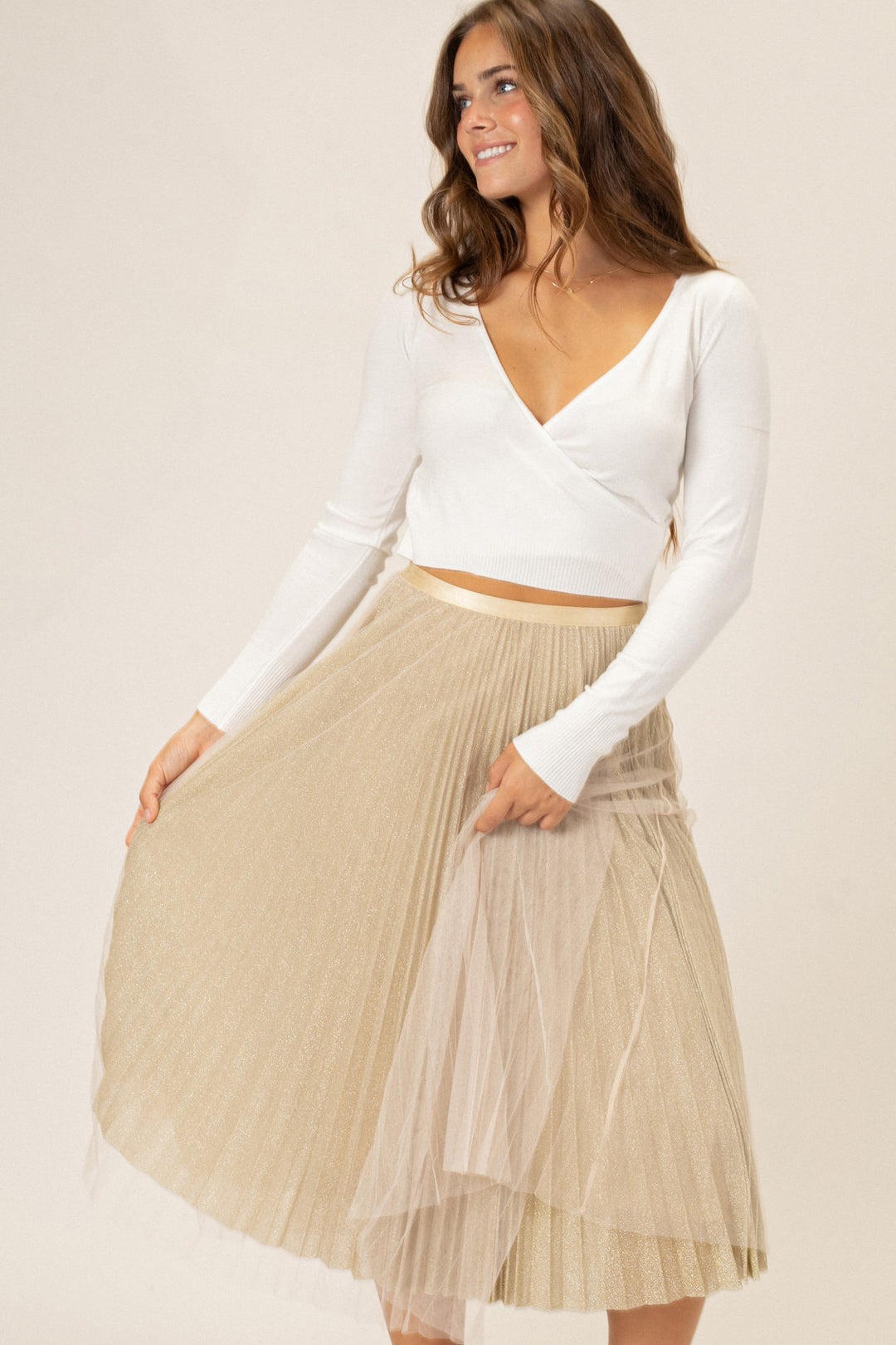 Elegant long skirt in a pleated gold-toned design, worn with a white cropped sweater for a chic layered look.