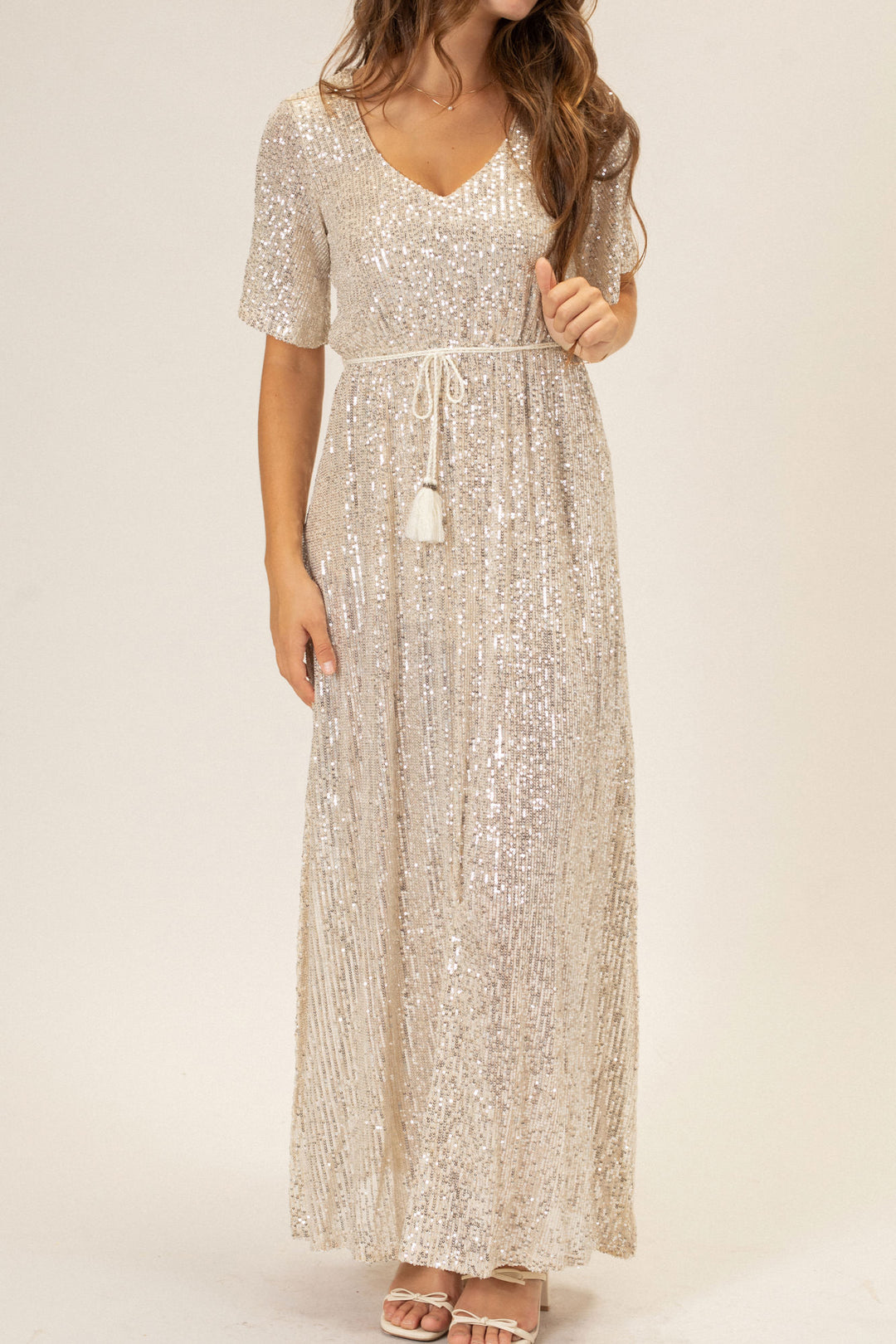 A long, sleeveless dress in a shimmering silver sequin fabric, with a V-neckline, a belted waistline, and a flowing maxi-length skirt.